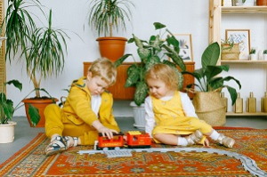 Dve deti v žltých šatách sa hrajú doma v obývačke na koberci s vláčikom